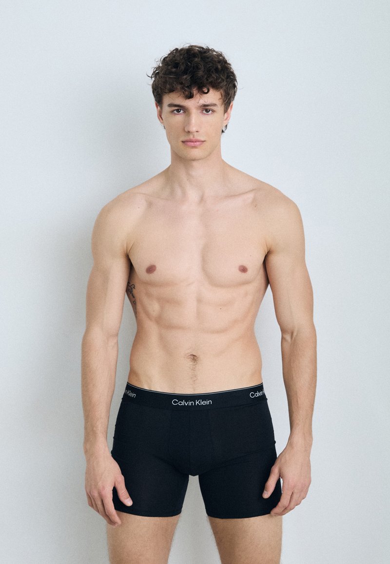 Schwarze Baumwoll-Boxershorts mit einem breiten elastischen Bund, der das "Calvin Klein"-Logo trägt; das Modell zeigt eine neutrale Haltung vor einem hellen Hintergrund.