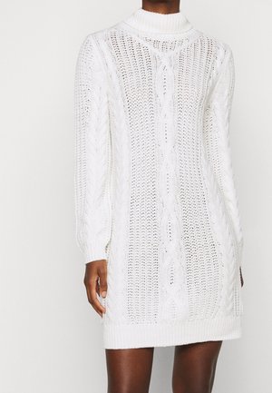 Person trägt ein weißes Kabelstrick-Turtleneck-Strickkleid mit langen Ärmeln und einem gerippten Saum, steht vor einem schlichten Hintergrund.