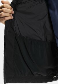 Interior de chaqueta negra con tejido suave y un bolsillo de malla. Las características incluyen un cierre con cremallera, dobladillo elástico y un diseño aerodinámico.