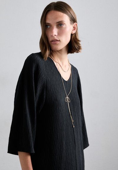 Vestido negro texturizado con escote en V y mangas de tres cuartos. Collar largo y delicado con un acento de lazo complementa el diseño.