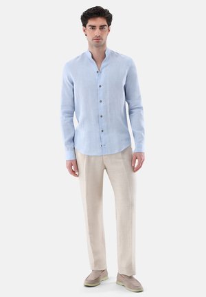 Mann trägt hellblaues Hemd mit Stehkragen, beige Hose und taupefarbene Slipper, steht vor weißem Hintergrund.