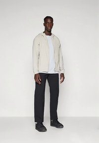 Lys beige lett jakke med hette, kombinert med en grå t-skjorte, svarte bukser med rett ben og svarte sneakers.