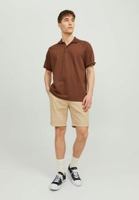 Polo shirt rayé marron, à manches courtes, associé à un short beige. Des baskets noir et blanc avec des chaussettes blanches complètent la tenue décontractée.