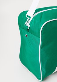 Bolso de nailon verde con detalles en blanco, forma rectangular, cierre superior con cremallera y un pequeño logotipo en la parte delantera. Incluye una correa ajustable.