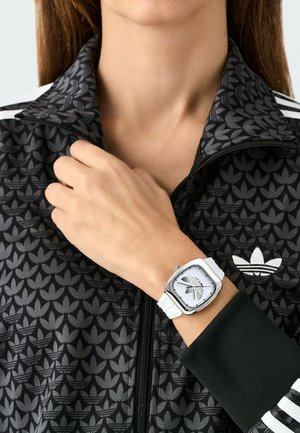 Persoană purtând o jachetă Adidas neagră cu model și dungi albe, arătând un ceas de mână cu cadran pătrat alb și ace negre pe încheietura mâinii stângi.