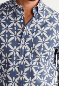 Camicia a maniche corte con bottoni in denim blu con motivo floreale bianco. Presenta un colletto e una tasca sul lato sinistro del petto, texture liscia.