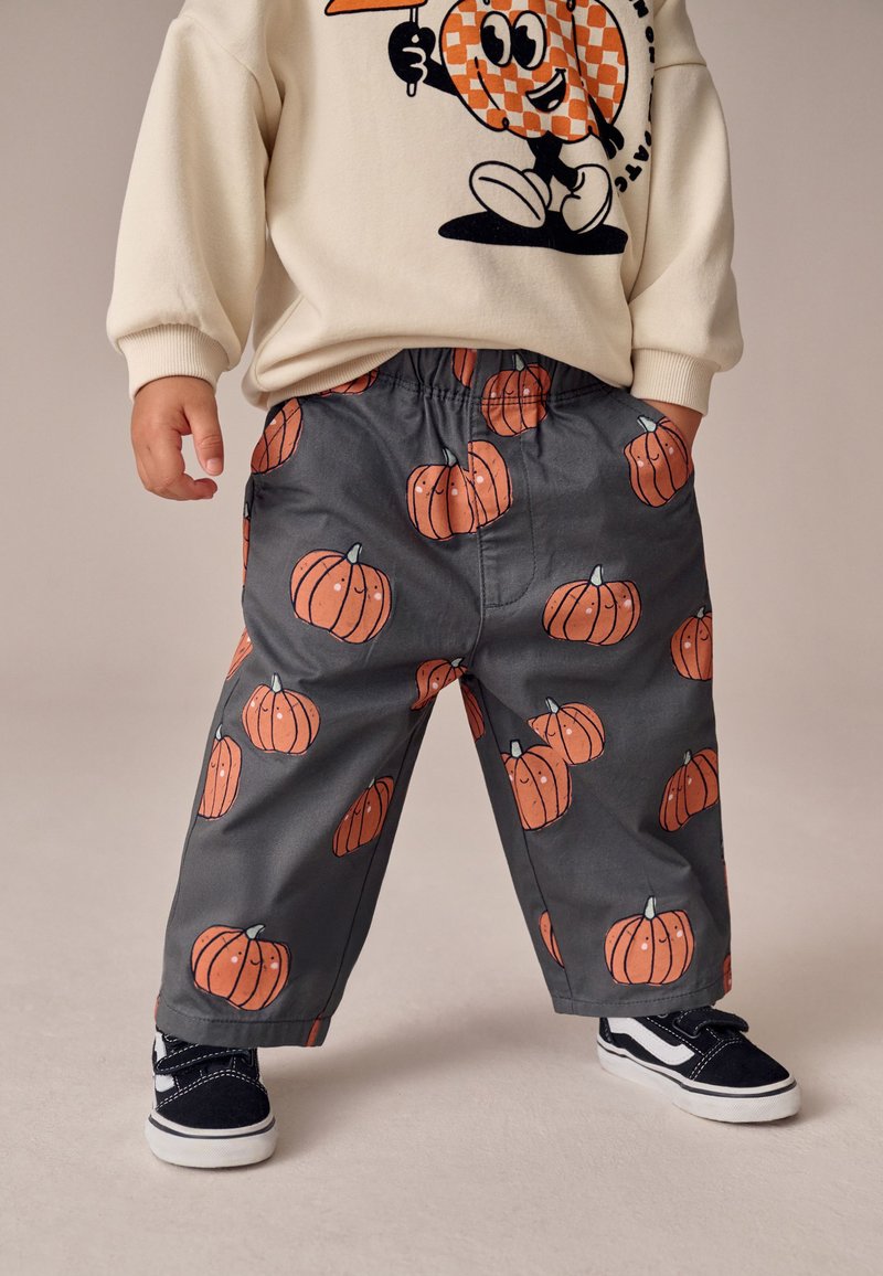 Pantalon gris avec imprimés de citrouilles orange vif, taille élastique et poches latérales, associé à des baskets noires et un sweat crème.