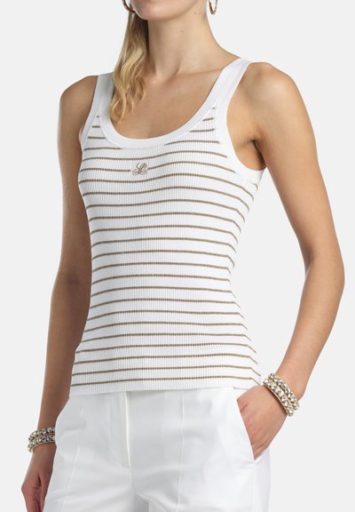 Femme portant un débardeur blanc côtelé avec de fines rayures horizontales marron et un logo "L" doré, assorti à un pantalon blanc et des bracelets.