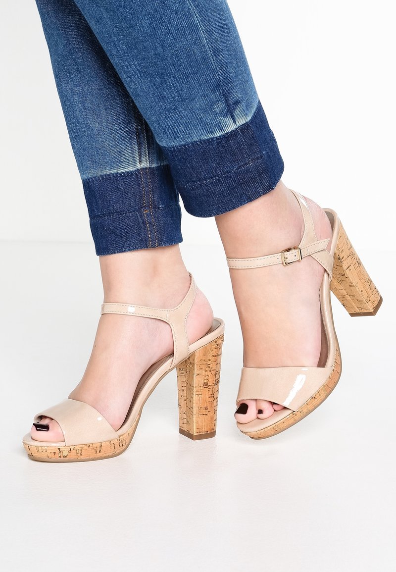 Pieds en sandales à talons hauts en cuir verni beige avec talons épais en liège et brides à la cheville, associées à un jean bleu à revers.