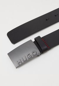 Zwarte leren riem met een matte grijze metalen gesp gegraveerd met "HUGO." Heeft rode stiksels op de lus van de riem. Glad textuur.