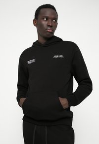 Moleton preto com um bolso frontal, apresentando gráficos em texto branco no lado esquerdo do peito e no ombro. Tecido suave com um corte descontraído.