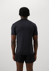 Herren passgenaues schwarzes kurzärmeliges Sporthemd. Glatte Textur, Rundhalsausschnitt und dezente Nähte. Getragen mit schwarzen Shorts, Rückansicht gezeigt.
