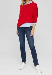 Kvinde iført en rød ribbet sweater over en lys blå skjorte, mørkeblå skinny jeans og hvide sneakers, stående mod en ensfarvet baggrund.