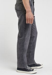 Man som bär urblekta svarta raka jeans och svarta lågt skurna sneakers med vita snören och sulor, stående på sidan mot vit bakgrund.