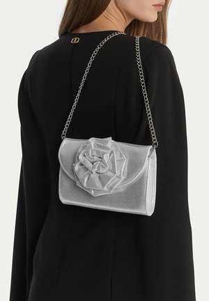 Donna con blazer nero che tiene una borsa a mano argentata con un grande fiore di stoffa e una tracolla a catena.