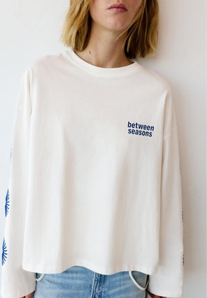 Mladá osoba nosiaca bielu košeľu s dlhým rukávom s modrým nápisom "between seasons" a modrými slnečnými vzormi na rukávoch, skombinovanú so svetlomodrými džínami.