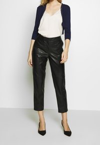 Svart läderbyxor med cropped-design och slät yta, matchat med en krämfärgad blus med v-ringning och en marinblå kavaj med spetsdetaljer. Svarta klackar.