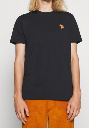 Osoba nosząca czarny t-shirt z krótkim rękawem z małym haftowanym pomarańczowym logo konia na piersi oraz pomarańczowe spodnie z kordurowej tkaniny.