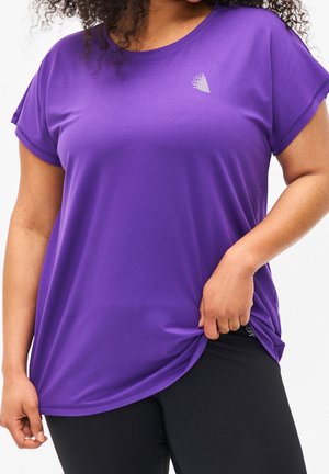 Kobieta ubrana w luźną fioletową koszulkę z krótkim rękawem z małym białym logo na piersi, trzymająca brzeg koszulki jedną ręką.