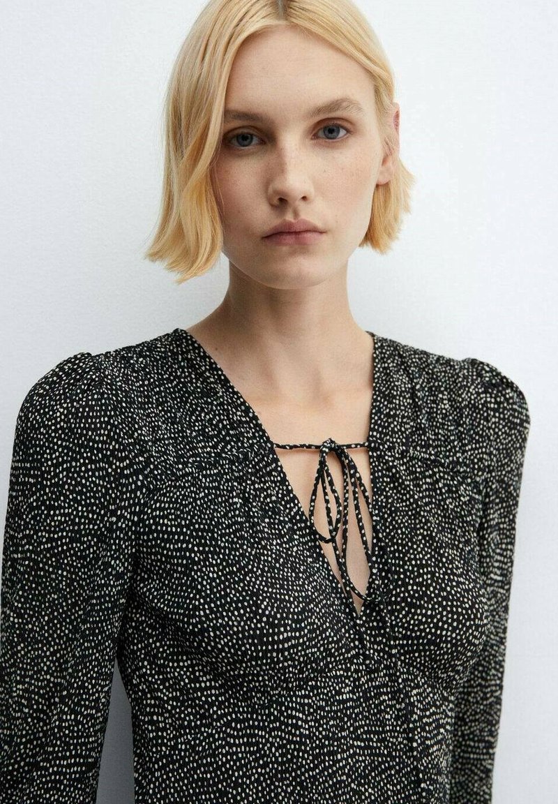 Vestito nero con motivo a pois bianchi, scollatura a V, dettaglio con cordino, maniche lunghe e tessuto testurizzato con leggero lucido.