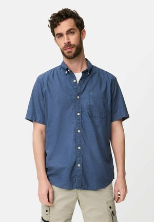 Mand med krøllet hår iført en blå kortærmet skjorte med knapper og beige cargobukser, stående foran en ensfarvet hvid baggrund.