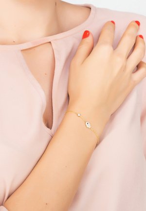 Gold bracelet with a hamsa design, featuring clear and blue circular accents, worn on a light-skinned wrist with coral nail polish.