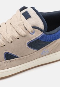 Sneaker in suede e mesh beige e navy, con lacci beige, un emblema blu e una suola in gomma bianca con finitura texturizzata.