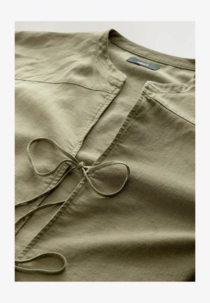 Blusa verde oliva confeccionada con tela suave, con un escote redondo y cierre frontal con lazo. Textura lisa con detalles mínimos.