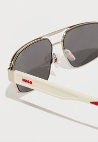 Okulary przeciwsłoneczne z złotą metalową oprawką, ciemnymi soczewkami i białymi zausznikami z czerwonym logo "HUGO". Projekt ma kątowe kształty i poduszki nosowe.