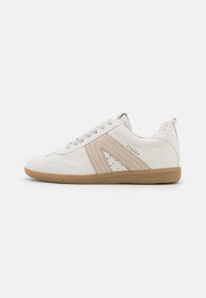 Zapatilla blanca de ante con detalle beige y suela de goma marrón texturizada. Presenta detalles perforados, diseño con cordones y tira para tirar.
