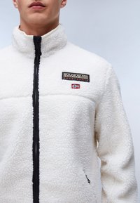 Weiße Fleecejacke mit hohem Kragen, Reißverschluss und schwarzen Akzenten. Enthält ein gesticktes Logo und ein kleines Detail der norwegischen Flagge.