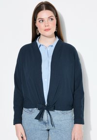 Cardigan cropped blu navy con chiusura anteriore a laccio, realizzato in un tessuto morbido. Abbinato a una camicia button-down azzurra e jeans in denim.