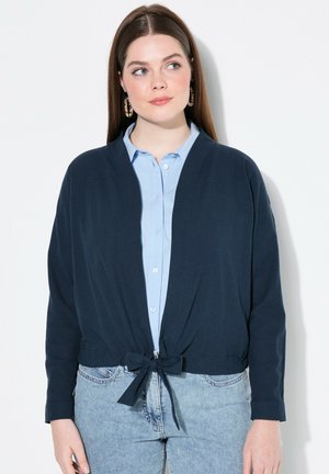 Cárdigan corto azul marino con cierre de lazo, hecho de una tela suave. Combinado con una camisa de botones azul claro y jeans de mezclilla.