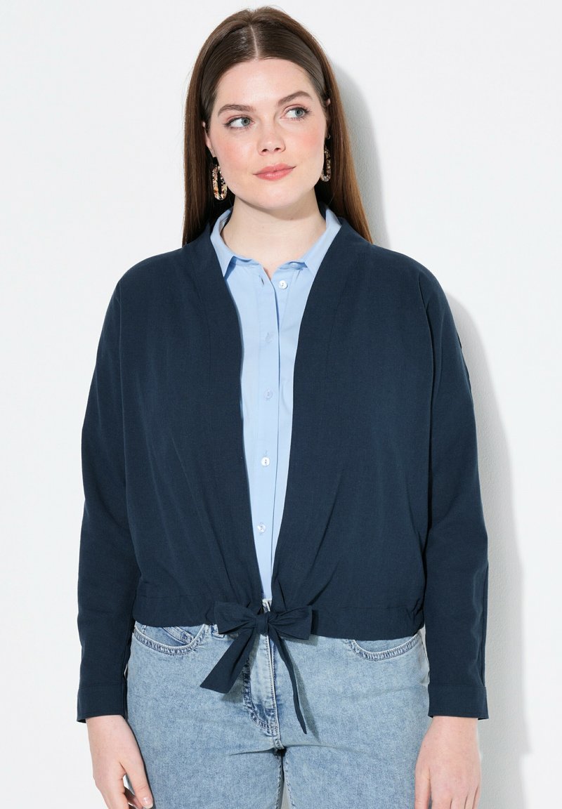 Cardigan cropped blu navy con chiusura anteriore a laccio, realizzato in un tessuto morbido. Abbinato a una camicia button-down azzurra e jeans in denim.
