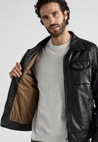 Zwarte leren jas met ritssluiting, twee voorkeurzakken en een bruine voering. Draagt over een lichtgrijze trui.