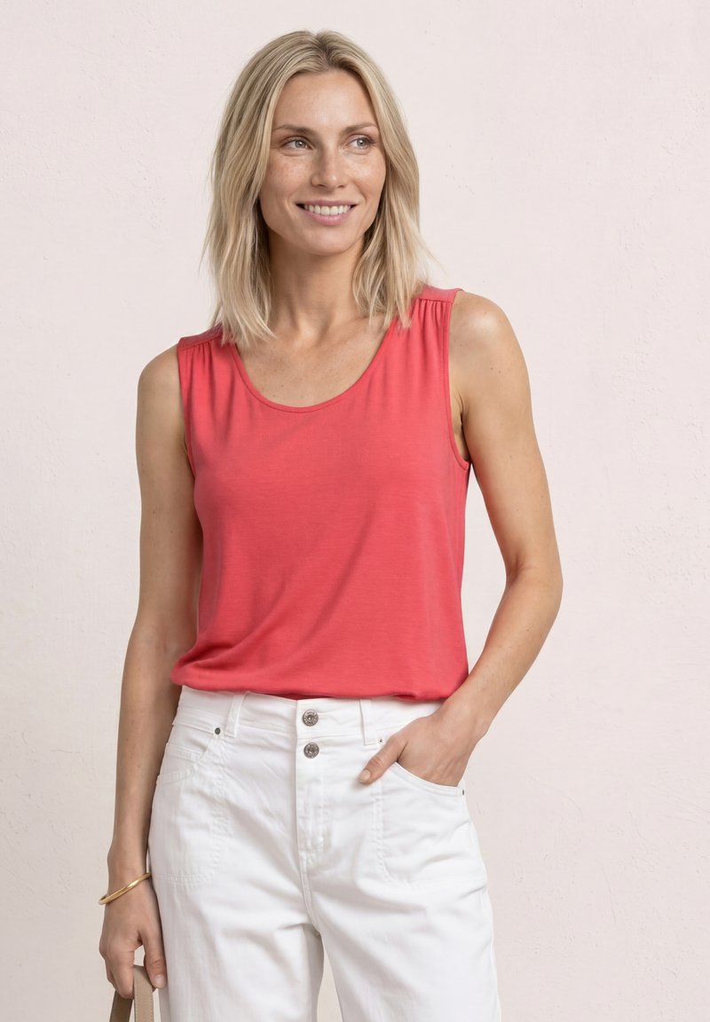 Femme blonde souriante, portant un haut corail sans manches et un pantalon blanc taille haute, debout avec une main dans la poche sur un fond pâle.