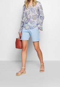 Un short bleu clair et un blouse à motif paisley multicolore, avec des sandales espadrilles beiges et un petit sac à main rouge.