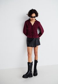 Burgunderfarbendes, gerüschtes Button-Up-Hemd, schwarzer Minirock aus Kunstleder und schwarze Schnürstiefel in kniehoher Form mit klobigen Sohlen. Eine Sonnenbrille rundet den Look ab.