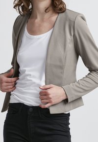 Person som justerar en ljusgrå kavaj över en vit skjorta, bärande svarta byxor, framför en enkel ljus bakgrund.