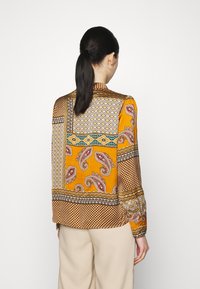Blouse à manches longues présentant un motif paisley en orange, bleu et crème avec des accents géométriques. Fabriquée en tissu lisse et léger.