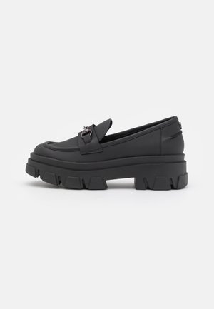 Mocassins noirs en matériau synthétique lisse avec une semelle en caoutchouc épaisse. Présente une sangle décorative et des accessoires en métal poli.