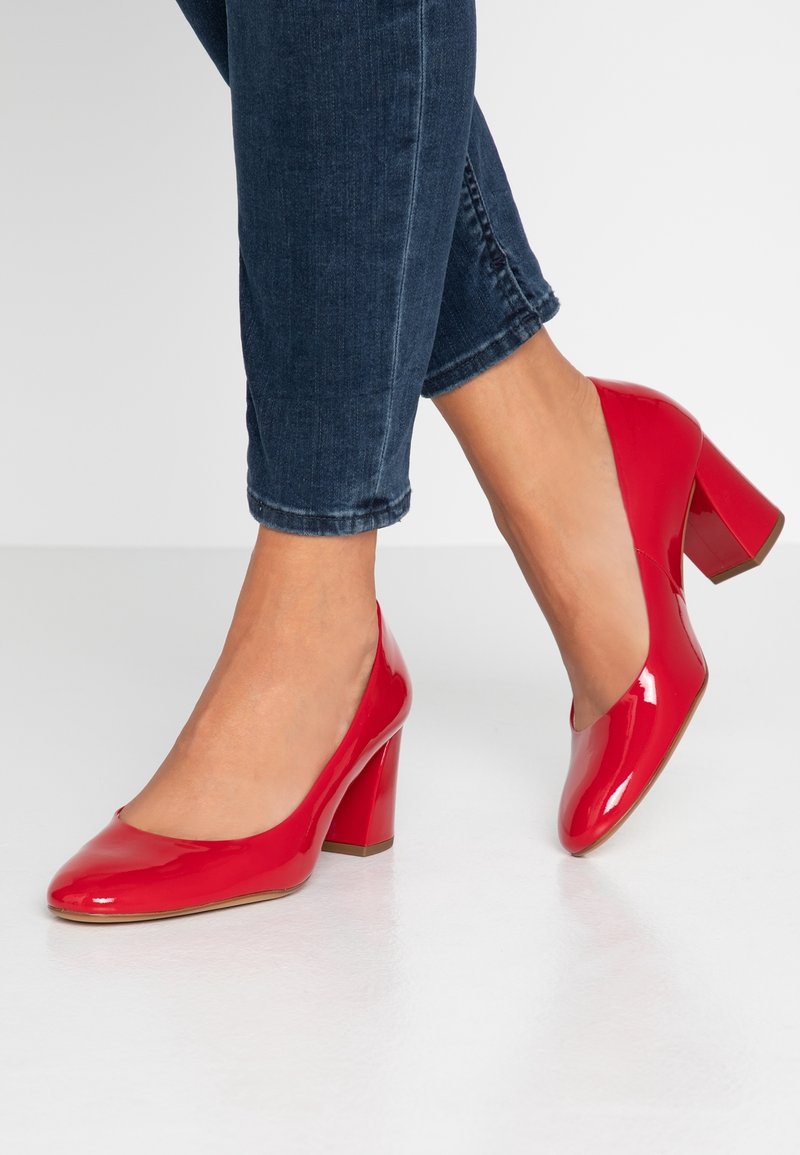 Chaussures à talons hauts en cuir verni rouge avec un bout rond et un talon épais, portées avec un jean court bleu foncé sur un fond blanc.