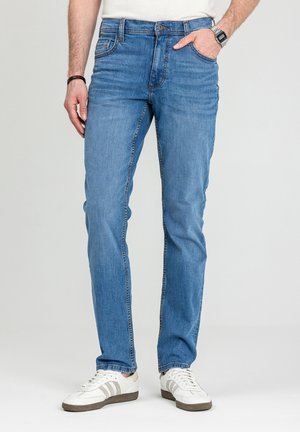 Mężczyzna w jasnoniebieskich dopasowanych dżinsach, białych trampkach, białej koszulce, czarnej opasce na nadgarstek i zegarku cyfrowym, z jedną ręką w kieszeni.