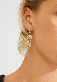 Gouden oorbel met een circulaire met kristallen versierde hanger en hangende bedels, waaronder parels en een roze steen, aan het oor van een vrouw.