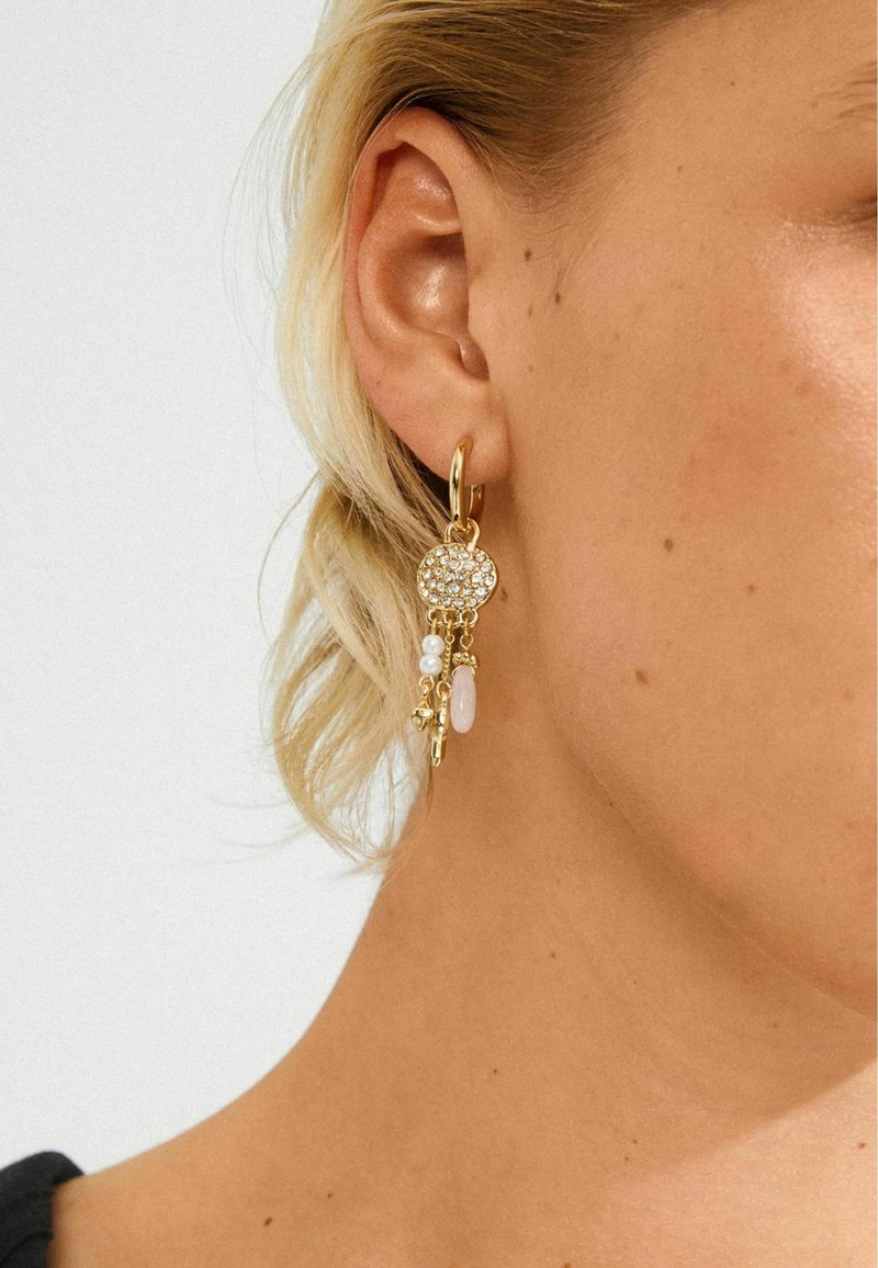 Gouden oorbel met een circulaire met kristallen versierde hanger en hangende bedels, waaronder parels en een roze steen, aan het oor van een vrouw.