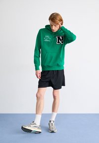 Green hooded sweatshirt with front pocket, embroidered logo, paired with black shorts and running shoes. Model in an indoor setting.