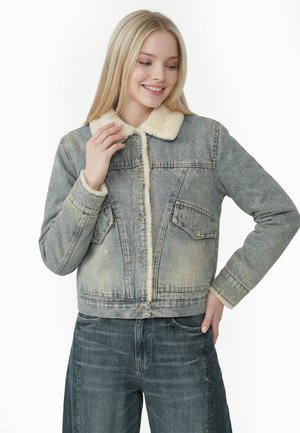 Ung kvinde smiler, iført falmet grå denimjakke med beige fåreskindskrage og -manchetter, kombineret med mørkegrå jeans, hånd på hoften.