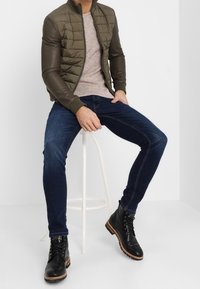 Olivgrön vadderad jacka med bruna lädersläva, grå stickad tröja, mörkblå jeans och svarta snörkängor på en vit pall.