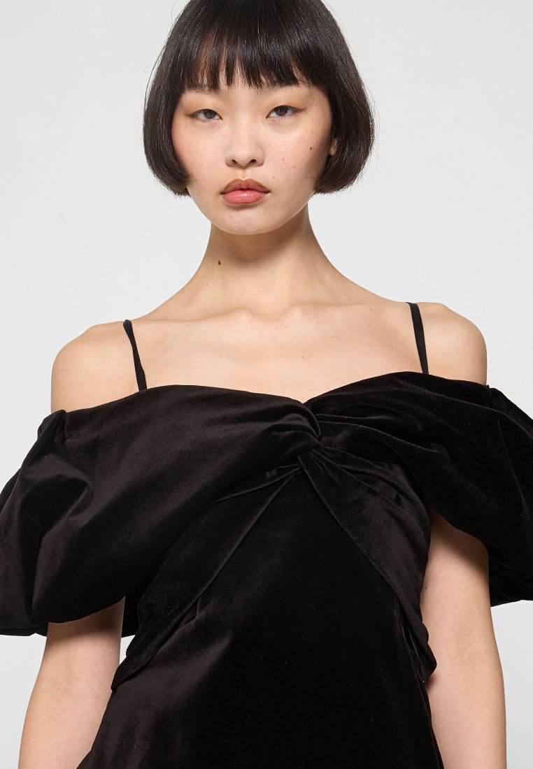 Robe en velours noir avec des manches bouffantes dénudées, un détail frontal torsadé et de fines bretelles noires. Texture lisse et drapé élégant.