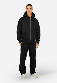 Casaco com capuz preto com fecho de correr frontal, bolsos frontais, punhos canelados e calças combinando. Feito de tecido macio, com um pequeno logótipo no peito.